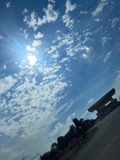 The image depicts a bright, sunny sky with scattered white cumulus clouds, suggesting fair weather. The sun is visible, casting strong rays and creating a lens flare effect, indicating it is likely daytime, possibly mid-morning or early afternoon. A black wire stretches diagonally across the sky, suggesting the presence of overhead electrical lines or communication cables.
In the upper right quadrant of the image, a portion of a road or paved area is visible, along with some trees and lampposts, hinting at an outdoor, possibly urban or suburban, setting. A yellow structure, possibly part of a building or signage, is also present. A car is visible in the distance.
The overall scene is dominated by the vast expanse of the sky, conveying a sense of openness and natural beauty. There is no clear activity or event taking place, and no people are visible. The rotation of the image suggests it was taken with the camera held at an angle.
No discernible text is visible in the image. The location context of Donskoe, Russia, places this scene within that geographic area.