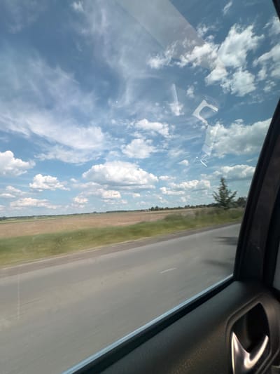 The image displays a rural landscape near Markivka, Ukraine, observed from the interior of a moving vehicle, specifically through a passenger-side window. The upper two-thirds of the scene feature a bright blue sky populated by numerous white cumulus clouds and scattered wispy cirrus clouds. Reflections of the sky and interior elements are visible on the window glass, particularly in the upper right quadrant.

Below the sky, a two-lane asphalt road extends horizontally across the middle to lower sections of the image, showing faint white dashed lines in the distance and suggesting forward motion due to slight blur in the foreground. Adjacent to the road, extensive flat agricultural fields are visible; some sections appear as tilled, brownish soil, while others are covered with green vegetation. A distant treeline defines the horizon, with barely discernible structures visible beyond it. A single evergreen tree, possibly a pine or fir, stands closer to the roadside on the right side of the landscape, exhibiting subtle motion blur.

The lower right portion of the image reveals a segment of the vehicle's dark gray, textured interior door panel, including a reflective, metallic door handle and a narrow silver-colored window trim. No individuals or animals are present in the captured environment. The scene is illuminated by bright daylight.