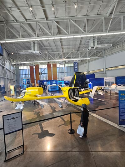 The image depicts a large indoor exhibition space with a high ceiling supported by exposed white metal trusses and ventilation systems. The floor is a polished concrete surface. The central focus is a bright yellow, futuristic aircraft with an open cockpit. This aircraft, identified by the "cora" logo, is positioned prominently in the foreground. To the right of the yellow aircraft, a white aircraft is partially visible.
Several large banners hang from the ceiling and walls. One banner, labeled "PERC", is visible in the upper left and center portions of the image. Other banners in the background display text related to "Boeing", "Aerospace", and "Education Lab".
A person, viewed from behind, is standing to the right of the yellow aircraft, holding a white shopping bag. The person is wearing dark clothing and a blue baseball cap. The aircraft's shadow is cast on the floor, along with the shadow of a standing sign to the left. This sign, labeled "GEN 05", displays an image of an aircraft and text.
The overall impression is that of a technology or aviation exhibition. This scene is located at the Boeing Future of Flight facility in Mukilteo, Washington, United States.