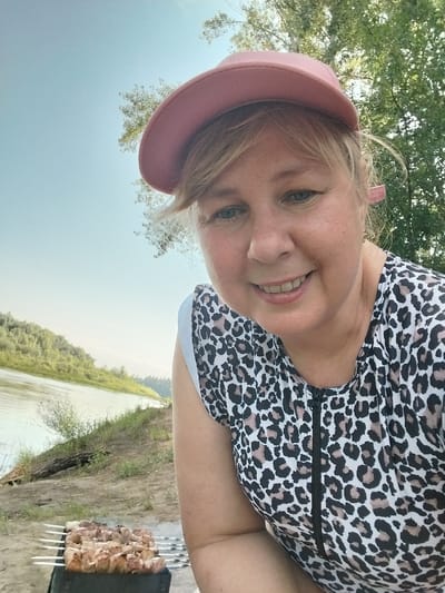 A close-up, selfie-style outdoor portrait shows a Caucasian woman with blonde hair and blue eyes wearing a pink baseball cap and a leopard-print top with a zipper. She is smiling and looking at the camera. In the background, there is a body of water, a grassy embankment, trees, and a blue sky. To the left of the woman, a charcoal grill with skewers of raw meat is visible. The scene appears to be a picnic or barbecue near a river. The location is the city of Chkalov, Russia.