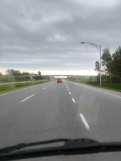 The image depicts a highway scene captured from the perspective of a vehicle's windshield. A red car is visible in the distance, driving on the highway. An overpass stretches across the highway ahead, with trees lining the sides of the road and the surrounding landscape. The sky is overcast with gray clouds. The scene is set in Rigaud, Canada.