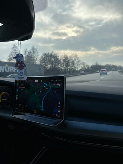 The image presents an interior view from a moving vehicle on a multi-lane highway, identified as the A35, observed near La Wantzenau, France. The central infotainment screen displays a navigation map indicating a route towards Schluchthal, Germany, with the current time as 15:37 and an interior temperature of 22.0 degrees Celsius.
A dark dashboard fills the lower frame. A cartoon-style air freshener, depicting a figure on a spring with a red shirt, cap, and "hopser" text, hangs from the rearview mirror. This air freshener partially obscures a large truck on the left side of the road, which bears a visible "WÜRTH" logo. The windshield exhibits fine water droplets. Ahead on the highway, a white sedan proceeds in the right lane, with other distant vehicles visible. The road is flanked by bare deciduous trees under an overcast sky showing some filtered light. No individuals are visible in the image.