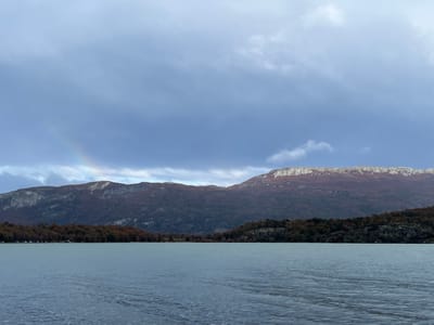 The image shows a landscape in Lapataia, Argentina, featuring a large body of water in the foreground, reflecting the overcast sky. The water surface has ripples, indicating movement or wind. In the middle ground, there is a line of trees with reddish-brown foliage, typical of autumn. Beyond the trees, there are mountains, one of which has a lighter, possibly snow-covered, ridge at its peak. The sky is filled with dark, heavy clouds, and a faint rainbow is visible in the upper left portion of the image.