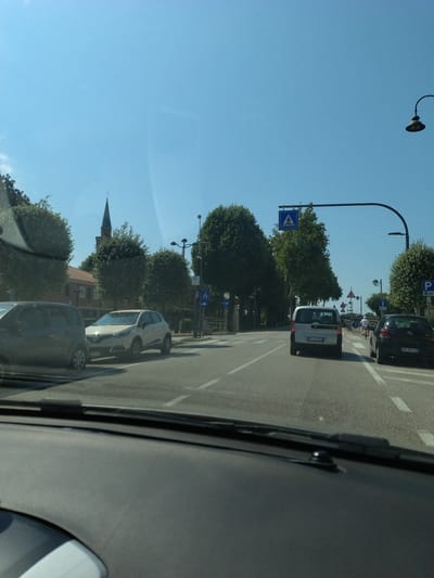 The image depicts a street scene in San Stino di Livenza, Italy, on a clear, sunny day. The perspective is from inside a vehicle, looking forward through the windshield. In the foreground, the dashboard of the car is visible.
On the street, there are several cars. A dark-colored car is partially visible on the left. In the center, a white Renault Captur is parked. Further down the road, a silver Peugeot is driving away from the camera. To the right of the silver Peugeot, a black car is also driving away.
A cyclist wearing a white shirt is visible on the right side of the road. There are pedestrian crossing signs with blue backgrounds and white silhouettes of people walking, indicating designated crossing areas. Traffic lights and street lamps are present.
In the background, tall green trees line the sides of the road. A red-brick building with multiple windows is visible behind some trees on the left. Dominating the skyline in the background is the spire of a church, characteristic of many Italian towns. The sky is a clear, bright blue. The road appears to be a multi-lane street with clear lane markings.