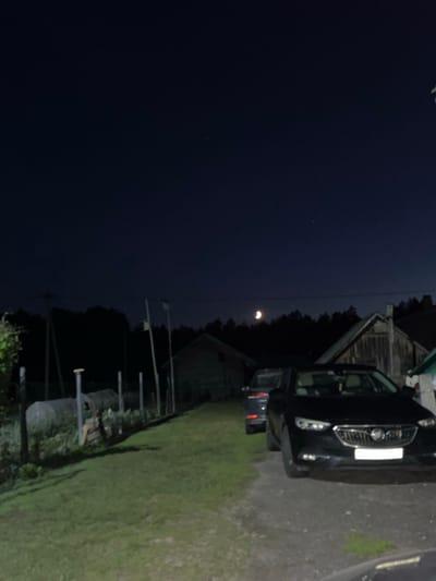 The image depicts a nighttime scene in the Miasotski Village Council, Belarus. In the foreground, a dark-colored sedan is parked on a gravel surface. Behind it, another vehicle, possibly an SUV, is also parked. Several wooden structures, consistent with rural dwellings or outbuildings, are visible in the midground. The environment appears to be a rural or semi-rural area, with trees forming a dark silhouette against the night sky. A crescent moon is visible in the upper portion of the sky. A grassy area and some fencing are present to the left of the parked vehicles. The overall lighting suggests it is nighttime, with the moon and possibly ambient light from the buildings illuminating the scene.