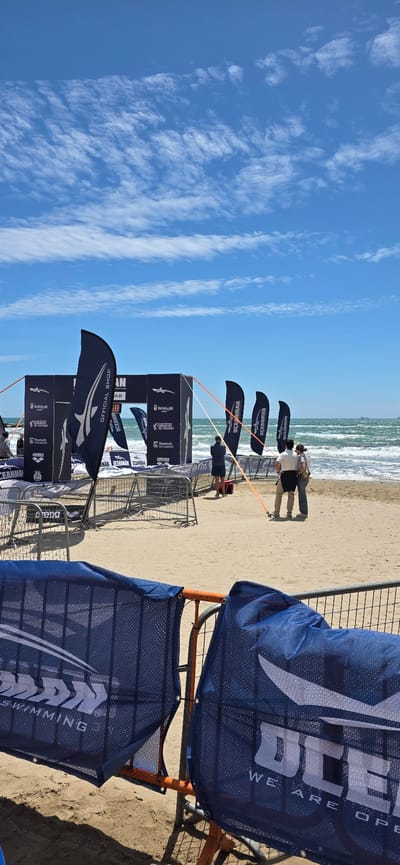 Here is a description of the image:
The image captures a beach scene in Benicàssim, Spain, with a view of the ocean on the horizon. The sky is clear with some wispy clouds.
In the foreground, there are barriers and flags set up. The flags are dark blue and display the logos of several entities, including "OCEANMAN", "Benicassim", "arena", and "Diputació de Castelló". The banners have an orange frame. Several people are standing on the beach further away in a huddle, their backs to the camera, seeming to look out towards the sea. The sand is light brown. The surf is relatively calm with small waves visible.