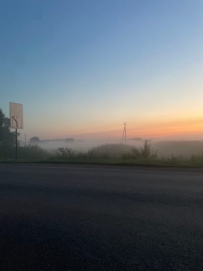 The image shows an early morning scene on a road in Uba, Russia. The sky is clear blue at the top, fading to a soft orange and yellow gradient towards the horizon, indicating sunrise. A thin layer of fog covers the distant fields, partially obscuring trees and a few scattered houses. A power line runs across the middle ground, supported by a single wooden pole. In the foreground, the dark asphalt of the road stretches across the bottom of the frame, with a white line marking the edge. To the left, a white traffic sign with a diamond shape and a downward-pointing arrow is mounted on a pole. There are no people visible in the image. The overall impression is one of a tranquil, rural landscape at dawn.