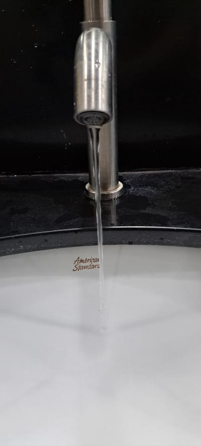 The image shows a close-up, vertical shot of a chrome faucet in operation, with a stream of water flowing into a white sink. The faucet is positioned in the center of the frame, and its metallic surface reflects light. Below the faucet, the white sink is visible, with the "American Standard" logo imprinted in brown on its surface. The background consists of a dark, possibly black, countertop or backsplash. Water droplets are present on the dark surface to the left of the faucet. The scene appears to be indoors, likely in a bathroom or kitchen. There is no indication of the specific location within Mandaluyong City, Philippines, beyond the context provided.