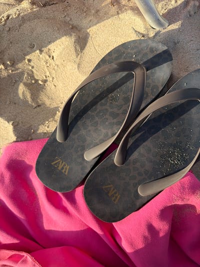 Two dark gray flip-flops with a subtle leopard print pattern are laid on a bright pink beach towel. The flip-flops are positioned on a sandy surface, with the towel beneath them. One flip-flop is positioned slightly above and to the left of the other. The right flip-flop has "ZARA" imprinted in gold lettering on its sole. Sand particles are visible on the surface of the flip-flops and the towel. In the background, a portion of a light-colored plastic object, possibly a chair leg or a similar beach accessory, is visible on the sand. The lighting suggests a sunny day, with distinct shadows cast by the flip-flops and the plastic object. The scene appears to be set on a beach in Urbanització Cales I Talaies, Spain.