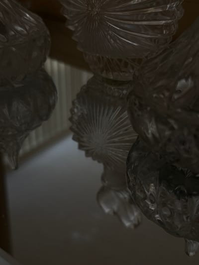 The image displays a close-up, upward-angled view of multiple transparent, intricately patterned glass objects, likely components of a chandelier or decorative fixture. At least five distinct glass elements are visible, each exhibiting varied patterns including radial "sunburst" designs, ribbed textures, and faceted surfaces. The ambient lighting renders the scene in shades of gray, with light interacting with the glass to produce reflections, refractions, and highlights that emphasize the material's transparency and the patterns. The objects are suspended above a dark, highly reflective horizontal surface, which shows blurred reflections of the glass. The background is significantly out of focus, revealing vertical striations consistent with window blinds or wall panels, and a horizontal dark wooden element, possibly a beam or ceiling fixture. No individuals are present. The scene is an indoor environment located in Hallandale Beach, United States.