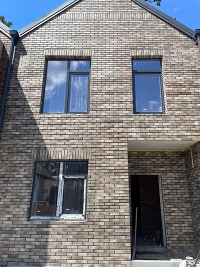 This is a full-frame, eye-level, exterior photograph of a multi-story building's brick facade under a bright, clear blue sky. The building appears to be under construction or recently renovated, as evidenced by the protective plastic sheeting on one of the windows and an open doorway revealing unfinished interior brickwork. The facade is constructed with variegated tan and brown bricks, laid in a running bond pattern with dark mortar.
There are three windows visible on the facade. The top left window is a rectangular pane of glass, reflecting the sky and clouds. The top right window is a narrower, vertical rectangular pane of glass, also reflecting the sky. The lower window is a larger, horizontal rectangular pane, partially obscured by a diagonal shadow cast from the left side of the building. This lower window also has protective plastic sheeting with branding, indicating it is new.
Below the lower window, an open doorway leads into a dark interior. A wooden-handled shovel or broom leans against the right side of the doorway. The threshold of the doorway is at ground level, with rough concrete steps visible inside. A yellow cable or pipe runs horizontally along the underside of a concrete overhang above the doorway. To the far left of the image, a downspout runs vertically down the side of an adjacent building, which has a red brick facade. The overall scene suggests a residential or commercial property in the city of Bucha, Ukraine, possibly undergoing renovations or repairs. No people are visible in the frame.