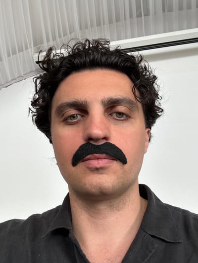 A man with curly dark hair and a fake black mustache is looking at the camera. He is wearing a dark t-shirt. The background is a white wall and a white blind. The image is taken from a low angle and is rotated 90 degrees clockwise. It is difficult to determine the time of day or weather from the image. The location is identified as a tourist boarding house "Klyazma Reservoir" in Russia, but there are no visual cues to confirm this. The man's expression is neutral. There is no visible text in the image.