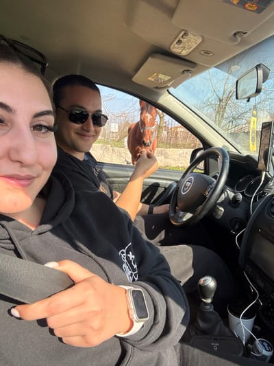 A selfie-style photograph captures two individuals inside a Dacia vehicle during daytime, in Zdravets, Bulgaria. In the foreground, positioned to the left, a female is visible from the chin up, featuring dark hair and long eyelashes. Her left hand, wearing a white smart watch with a sparkling bezel, rests on a black seatbelt, confirming she is buckled. She wears a black hoodie. Seated in the driver's seat, a male with short dark hair and sunglasses looks towards the camera with a slight smile. His right arm is extended through the open passenger window, holding a small object, appearing to feed a brown horse. The horse, wearing a light blue halter and exhibiting a white blaze on its forehead and white lower legs, has its head inside the car's window opening, interacting with the male. The car's interior details include a black steering wheel with the Dacia logo, a manual gear stick, a dashboard-mounted device holder, a rear-view mirror with a yellow air freshener, and a sun visor. Two white paper cups and charging cables are visible in the center console. Outside the car, a wire fence, sparse trees, and some greenery are visible under a bright, clear sky, with light-colored ground beneath.