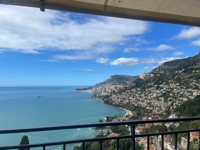 The image shows a panoramic view from a balcony overlooking the Mediterranean Sea and the town of Roquebrune-Cap-Martin, France. The balcony is situated on a hilltop, with a black metal railing separating the viewer from the scene below. The blue sky is dotted with white clouds. In the distance, the clear blue waters of the Mediterranean stretch out to the horizon. A small boat is visible in the water.  The coastline is lined with dense green vegetation, with buildings and houses visible on the slopes. The town of Roquebrune-Cap-Martin, with its white buildings and red tile roofs, extends along the hillside.  The scene is bathed in bright sunlight.