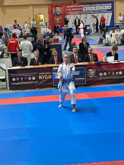 The image shows a karate competition in Demenshchina, Russia. A young woman wearing a white karate gi with a blue belt is performing a kata on a blue and red mat. A screen in the background reads "064 OHAN DAI," indicating the name of the kata. Spectators and judges are seated in the background. Banners displaying the event's name, "Всероссийские Соревнования по Каратэ" (All-Russian Karate Competition), and "Кубок Памяти Героя Советского Союза Полковника Григория Ивановича Бояринова" (Cup in Memory of Hero of the Soviet Union Colonel Grigory Ivanovich Boyarinov), are also visible.