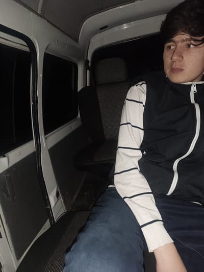 A young individual, likely male, is seated in the interior of what appears to be a compact utility vehicle or van. The person is positioned in the foreground, facing slightly away from the camera and looking towards the right side of the frame with a neutral, observant expression. They have dark, slightly tousled hair and light skin. A white earbud is visible in their right ear.
The individual is wearing a dark blue or black sleeveless vest with a white zipper running down the front and white trim along the edges. Underneath, they have a light-colored (possibly off-white or beige) long-sleeved shirt with thin, dark horizontal stripes. Dark blue jeans cover their legs.
The interior of the vehicle is utilitarian, with white metal or plastic panels forming the ceiling and side walls. The windows on the left side of the vehicle appear dark, suggesting it is nighttime or a very dim environment outside. The door on the left side is slightly ajar, revealing more darkness outside. The seating consists of dark grey or black fabric upholstery with a subtle patterned texture. A headrest is visible on the seat behind the person, and part of a seatbelt mechanism can be seen near it. The overall lighting inside the vehicle is dim, casting shadows but still illuminating the person's face. No text is visible in the image.