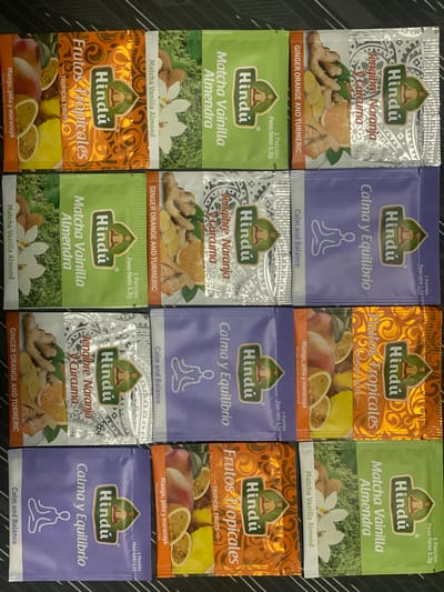 The image displays a collection of eight individual tea sachets arranged in two rows of four. The sachets are predominantly vibrant in color, with distinct designs and text indicating different flavors.

**Top Row (Left to Right):**
1.  **Orange Sachet:** Features a tropical fruit illustration with text in Spanish that translates to "Tropical Fruits." The brand logo "Hindú" is prominently displayed.
2.  **Green Sachet:** Displays matcha powder, almonds, and a flower. The text indicates "Matcha Vanilla Almond" in Spanish and English.
3.  **Orange and Ginger Sachet:** Shows slices of orange and ginger. The text reads "Ginger Orange and Turmeric" in English and Spanish.
4.  **Silver Sachet:** Features a pattern and the text "Jengibre Naranja y Curcuma" (Ginger Orange and Turmeric) in Spanish.

**Bottom Row (Left to Right):**
1.  **Orange Sachet:** Similar to the first in the top row, with "Frutos Tropicales" (Tropical Fruits) and fruit illustrations.
2.  **Purple Sachet:** Depicts a meditating figure with text "Calm and Balance" in English and "Calma y Equilibrio" in Spanish.
3.  **Orange and Ginger Sachet:** Similar to the third in the top row, with ginger and orange imagery.
4.  **Green Sachet:** Similar to the second in the top row, with matcha powder, almonds, and a flower.

The sachets are laid out on a dark, textured surface, likely a table or countertop. The lighting appears to be ambient, casting soft shadows. The perspective is slightly overhead, capturing the top surfaces of the sachets. The image is a still life, focusing solely on the product packaging. There are no people, animals, or any indication of a specific city or location, including Santa Marta, Colombia. The context is purely commercial, showcasing tea products.