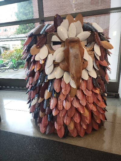 This image depicts a large, cylindrical, abstract sculpture, approximately human-height, situated indoors. The sculpture is composed of numerous overlapping, glossy, leaf-shaped tiles or scales in varied tones of maroon, rust, dark brown, dark blue, and cream. A central, vertically oriented, rough-hewn wooden element forms a prominent beak-like or facial feature on its upper portion. The sculpture stands on a polished light-grey floor, with a dark-grey carpet in the immediate foreground. Behind the sculpture, large, dark-framed windows offer a view of an outdoor urban setting in Seattle, United States. Visible outside are green trees, a multi-story building with light-colored facades, and a paved area where two green and white shared bicycles (Lime bikes) are parked.