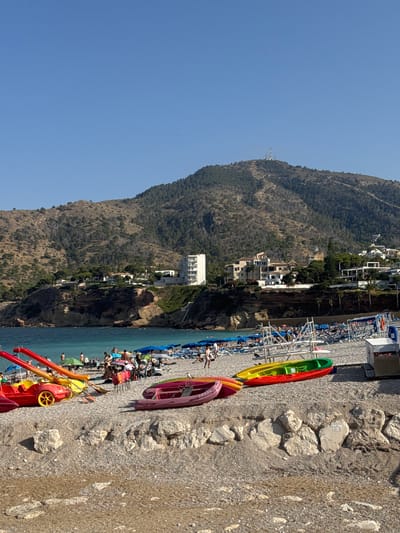 This is a photograph of a crowded beach on a sunny day, likely in the afternoon given the bright sunlight and long shadows. The scene is set against a backdrop of a steep, tree-covered mountain that rises directly from the coastline. Several white buildings, likely hotels or apartments, are nestled into the hillside.

In the foreground, the beach is covered in pebbles and sand, with a line of colorful kayaks and paddle boats resting on the shore. Some of these boats are red, yellow, green, and purple. Beyond the kayaks, a section of the beach is set up with rows of lounge chairs and umbrellas, many of which are occupied by people. The water is a clear, bright blue and appears calm.

Numerous people are visible on the beach, some relaxing on lounge chairs, others walking, and some swimming or wading in the shallow water. The atmosphere appears to be one of leisure and enjoyment. The image also captures a sign with some faint text, but it is unreadable. The overall impression is a vibrant Mediterranean beach scene with a dramatic natural landscape.