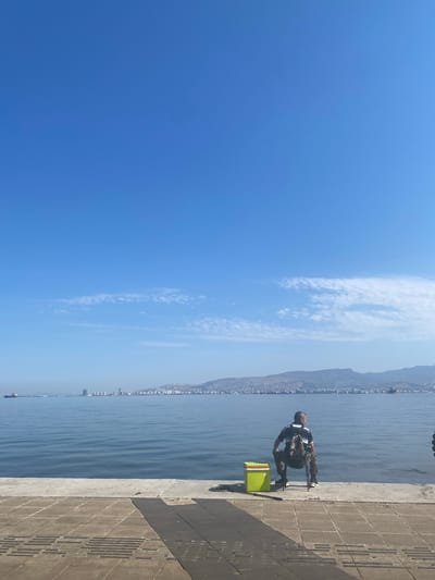 The image captures a man fishing from a concrete quay along the coast of İzmir, Turkey, on a clear, sunny day. The man, dressed in dark clothing and a light-colored vest, is seated on a small stool beside a green fishing box. He is looking out at the water.

The scene includes the sea, a cityscape in the distance at the base of a mountain range, and a bright blue sky with some wispy clouds. The quay is made of patterned concrete slabs.