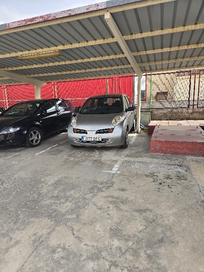 The image displays a partially covered outdoor parking area located in Valverde del Majano, Spain. Two passenger vehicles are parked on a concrete surface exhibiting cracks, discoloration, and worn white parking bay lines. In the foreground, a silver Nissan Micra (K12 generation) is positioned facing the viewer, displaying the Spanish license plate L 077 DFC. To its left, a black SEAT Leon (second generation) is partially visible, parked facing away from the viewer.
The parking area is sheltered by a metal canopy structure featuring grey corrugated roof panels supported by cream-colored metal beams and columns. A fluorescent light fixture is affixed to the underside of the canopy. The visible front edge of the canopy structure shows areas of peeling red paint and minor corrosion. In the background to the left of the vehicles, a corrugated red metal fence or wall extends horizontally. To the right, behind a metal support column, a light-colored building with a patterned facade is discernible, partially obscured by a brown metal security fence or railing. On the concrete surface in the right foreground, a low, rectangular reddish-brown structure, possibly a planter box or utility cover, is present. No individuals are depicted in the scene.