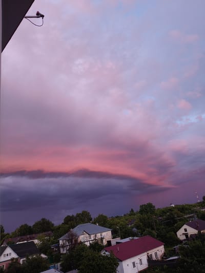 The image depicts a wide-angle view of a residential area at sunset, with a dramatic sky filled with pink and purple clouds. The foreground is dominated by the corner of a dark-colored structure, possibly a building or balcony, from which a security camera is mounted. Below the sky, a cluster of houses with various roof colors, including white, gray, and red, are visible nestled amongst dense green trees. Power lines crisscross the scene between the houses. The overall atmosphere is serene, illuminated by the soft, colorful light of the setting sun. The specific location is the city of Petrovsk, Russia. There are no people or animals visible in the image.