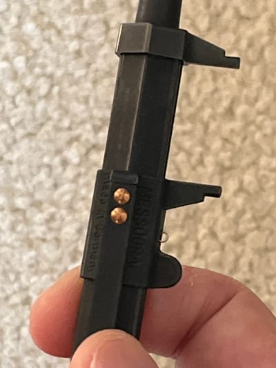 The image shows a close-up view of a dark-colored measuring tool, possibly a specialized caliper or gauge, held by a person's hand. The tool is oriented horizontally against a softly blurred, light-colored, textured background, which appears to be carpet or fabric.

The tool consists of a main rectangular bar and a parallel cylindrical rod. On the left, a fixed jaw-like component is visible. A sliding mechanism is present further right, featuring an upright, notched part. To its right, another sliding section includes a small, curved wire detail and a rounded protrusion. Two small, round, metallic studs, appearing coppery or brass-colored, are embedded in the tool's surface.

Visible text on the tool, appearing inverted, includes "MESSOGRAL" and "made in germany". A person's hand, with light skin, holds the right portion of the tool. No specific measurement or action is being performed, only the object being held. No explicit location cues are visible in the image, other than the general context of Middle Park, Australia.