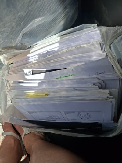 This is an eye-level, close-up shot of a stack of papers held within a clear plastic bag. The papers are predominantly white and appear to be official documents or forms, with some handwritten notes in blue and green ink. One document features a diagram of two human figures and text that includes "Devitnieku likums" (Law of Devitnieki), "Pieauguša cilvēka ķermeņa virsmas % sadalījums" (Adult human body surface % distribution), and a list of body parts with corresponding percentages. Another section of text appears to be related to "Zīdaiņa ķermeņa virsmas % sadalījums" (Infant human body surface % distribution), also with percentages. A yellow sticky note is attached to one of the papers. The setting is dimly lit, suggesting an indoor environment, possibly a storage area or a workshop, given the presence of other plastic-wrapped items in the background. Two pairs of human legs wearing white sandals are visible at the bottom right of the image, suggesting someone is holding or interacting with the stack of papers. The overall impression is one of documentation and organization, with a hint of an ongoing task or project.
