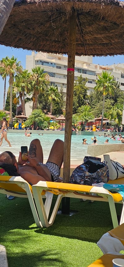 A group of people are enjoying a sunny day at a resort pool. The image shows a man relaxing on a yellow lounge chair, holding a smartphone. He is wearing patterned swim trunks. Beside him, a patterned beach bag and a white and blue bag are placed on the chair. The background reveals a pool filled with people swimming and wading, palm trees, and a multi-story building. The scene is set in Algeciras, Spain, under clear blue skies, indicating pleasant weather. The overall atmosphere appears relaxed and leisurely. The time of day is likely midday or early afternoon, given the bright sunlight casting shadows.
