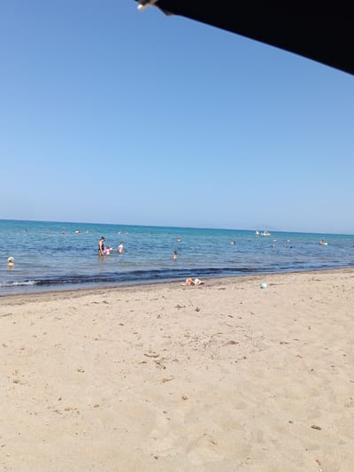 The image shows a sandy beach with people swimming and relaxing in the clear blue sea under a bright sky. The foreground is dominated by the sand, with a few scattered pieces of debris and footprints. In the middle ground, the water is calm with small waves lapping at the shore. Several people are visible in the water, some wading and others swimming. A few objects, including a ball and some beach toys, are present on the sand near the water's edge. The background shows the expanse of the sea stretching to the horizon, with a few boats and distant landmass. The scene appears to be a typical summer day at a coastal resort. This location is likely within the Municipal Unit of Larissos, Greece, known for its beaches.