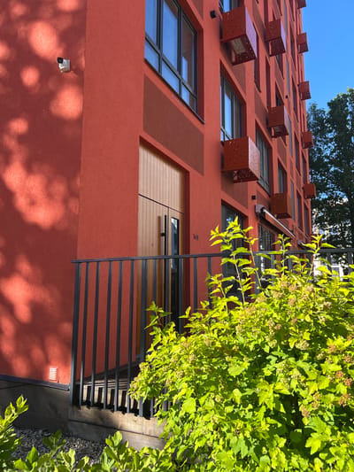 The media file displays a close-up, upward-facing view of a modern, red-walled building with numerous windows and distinctive, reddish-brown cube-like structures protruding from its facade. These structures, some with perforated designs, appear to be decorative or functional elements, possibly air conditioning units or unique architectural features. The building's texture is rough, suggesting stucco or a similar material.
Various windows of different sizes are visible, some reflecting a clear blue sky. On the lower right side of the frame, lush green foliage, likely from a tree or shrub, partially obscures the building, casting dappled shadows on the red wall. A black metal railing runs horizontally across the middle section of the building, hinting at balconies or a walkway. A security camera is mounted on the lower part of the wall.
The lighting suggests it is daytime, with bright sunlight creating strong contrasts and shadows. The clear blue sky indicates fair weather. The presence of a building with modern architectural elements, green vegetation, and the specific mention of Kaluga, Russia, provides context for the location. There is no visible activity of people, suggesting the image focuses on the architecture and its surroundings. A small, dark numeral "149" is visible on the red wall near the railing.