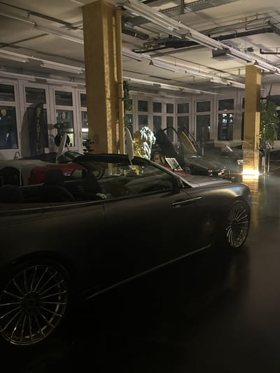 A dark, poorly lit photograph shows a car parked on a street, possibly in Wetzikon (ZH), Switzerland, although specific landmarks are not clearly identifiable. The car is a dark color, reflecting some ambient light. In the background, a building with multiple windows is visible, and architectural details suggest an urban setting. The overall scene appears quiet, possibly at night. The precise make and model of the car are not readily discernible.