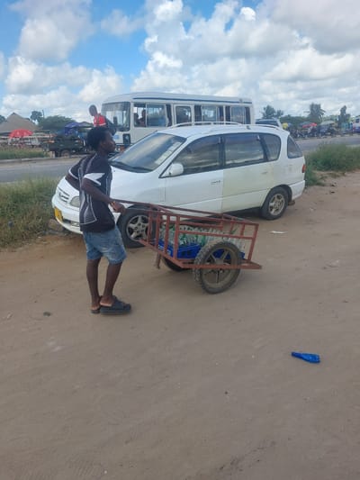 The image shows a man standing next to a white van with a trailer attached. The man is wearing a black and white t-shirt, blue shorts, and black sandals. He is looking towards the van. The trailer is made of red metal and contains several bottles of water. The van is parked on a dirt road next to a grassy area. In the background, there are other vehicles, people, and buildings, suggesting a busy roadside scene. The sky is cloudy. A blue plastic bag is visible on the ground to the right of the van. There is no visible text. The setting appears to be Picha Ya Ndege, Tanzania. The overall impression is that of a commercial or transportation hub.
