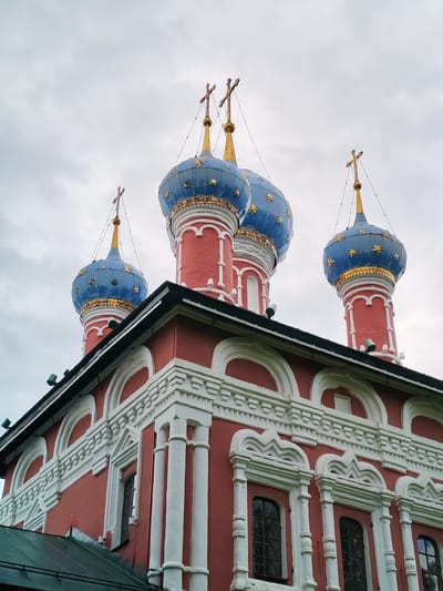 This is a low-angle, close-up, outdoor, eye-level shot of the cathedral domes and upper facade of a Russian Orthodox church in Selivanovo, Russia. The sky is overcast with faint clouds and numerous small dark specs that may be birds or debris. The domes are bulbous and painted bright blue, adorned with gold stars and gold trim. Each dome is topped with a gold cross. The body of the church visible is painted a vibrant red with intricate white architectural molding, featuring arched windows with decorative surrounds and columns. A dark green sloped roof is visible in the lower left corner. There are no people or animals present in the image. The lighting is diffused due to the cloudy sky.