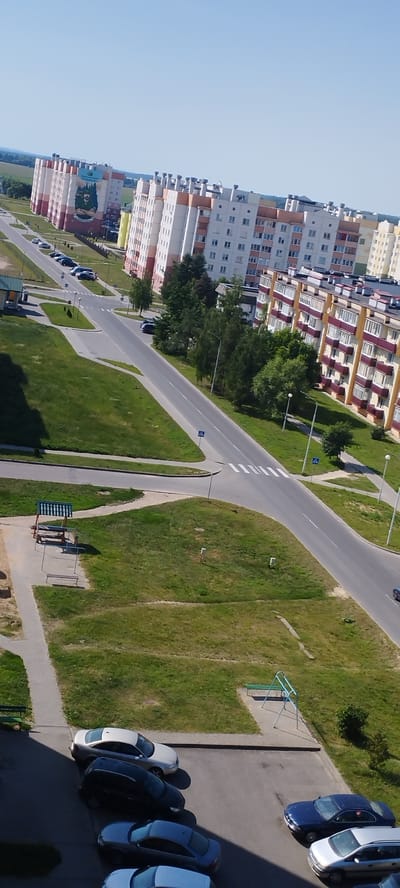An aerial, slightly tilted, eye-level long shot captures a sunny day in the city of Skidzieĺ, Belarus. The scene is dominated by modern, multi-story apartment buildings with light-colored facades and red accents, lining a wide, paved road. The buildings feature numerous windows and balconies. The road curves through a residential area with well-maintained green lawns, scattered trees, and a few parked cars. One building features a large mural depicting a man in a green hat. A pedestrian crossing is visible on the road. In the foreground, several cars are parked on a paved surface near the base of the buildings. Playground equipment, including a swing set, is visible on the grass. The sky is clear and blue.