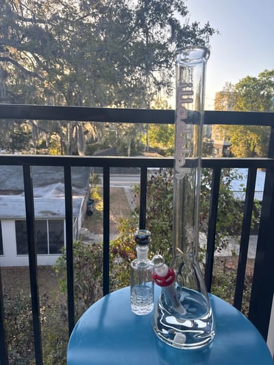 The image depicts a clear glass beaker-style water pipe, or bong, containing water, positioned on a round, blue outdoor table. To its left, a smaller clear glass ash catcher with a patterned base and a dark-colored bowl piece is also on the table. The water pipe features a vertical "Stundenglass" logo in white text on its neck and a red silicone grommet connecting its downstem. The scene is viewed from an elevated balcony, indicated by black metal railings in the foreground and midground. The background reveals an urban environment consistent with Gainesville, United States of America, characterized by large trees, likely live oaks, heavily draped with Spanish moss. Sunlight, suggesting either morning or late afternoon, illuminates the scene from the right, casting soft light on the trees and buildings. Below the balcony, rooftops of single-story residential buildings with light gray shingles and a screened-in porch are visible, alongside dense green foliage and a paved road or alleyway. In the distance, other multi-story buildings are observable. No individuals or active interactions are present in the frame.