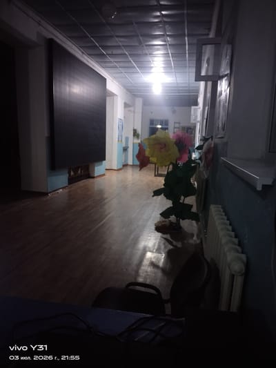 The image depicts a dimly lit interior hallway, possibly in a school or institutional building, characterized by a wooden floor, a suspended ceiling with visible panels and light fixtures, and a long, dark, rectangular display screen mounted on the wall. To the right, a collection of large, artificial flowers in vibrant yellow, pink, and red hues are arranged, some with green foliage, on the floor. Adjacent to the flowers is a white radiator and a dark blue wall. In the foreground, dark furniture, possibly chairs and a desk, are visible. The lower left corner of the image contains a watermark that reads "vivo Y31" and the date "03 июля. 2026 г., 21:55". The overall ambiance is quiet and still, with artificial lighting casting long shadows. The location is not specified as Moscow, Kazakhstan.