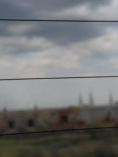 The image shows a blurred view through a dirty window or glass surface with horizontal dividers. The top portion of the image is dominated by a cloudy, gray sky. Below this, a horizontal black line cuts across the frame. The middle section is a hazy, muted view of what appears to be buildings with some orange and yellow accents, suggesting a possibly industrial or dilapidated area. Another horizontal black line is present below this section. The bottom of the image shows a blurred green and brown area, likely ground vegetation. The overall impression is one of looking out at a dull, overcast day over a somewhat neglected urban or semi-urban landscape. The location is specified as Приречный ауылдық округі, Kazakhstan.