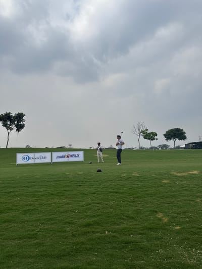 On a verdant golf course, two men are golfing under a cloudy sky. One man is in the midst of his swing. Nearby are two signs, one featuring "Ecuador Impulsa" and the other with "Diners Club". Sparse trees dot the landscape behind them, with buildings visible further in the background.