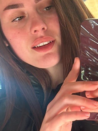 An image captures a close-up, angled selfie of a young adult female in Fowlis Easter, United Kingdom. She is positioned slightly off-center to the left, with her head tilted, revealing long, dark brown hair cascading over her shoulders. Her skin appears fair with a slight flush on the cheeks. She has defined eyebrows and greenish-brown eyes looking towards the upper left. Her lips are parted, showing her upper teeth and a hint of lower teeth, and are coated with a light, natural-toned lipstick. She is wearing a dark garment, likely a jacket, visible around her neck and shoulders. Her right hand (viewer's left) holds a dark, reflective smartphone with a patterned or textured case, positioned in the lower right portion of the frame. The background is indistinct and out of focus, suggesting an indoor setting with soft, natural lighting illuminating her face and hair.