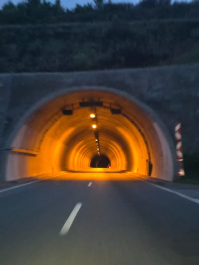 The image displays the entrance to a road tunnel, viewed from just outside, looking directly into its illuminated interior. The overall scene suggests it is either dusk or night, with the sky appearing dark blue or grey, and surrounding foliage to the right of the entrance being dark and indistinct.
On the right side of the tunnel's concrete entrance, a red and white striped warning barrier or sign is partially visible above. An asphalt road leads into the tunnel on the left, with a single, white dashed line painted on its surface.
Inside the tunnel, numerous bright, warm orange-yellow lights are arranged along the ceiling, casting a strong glow that illuminates the curved tunnel walls and floor. These lights create a clear sense of depth, drawing the viewer's eye along the tunnel's length. The tunnel appears to be made of concrete or a similar material and has a rounded, possibly semi-circular, cross-section.
No people, vehicles, or specific activities are depicted; the scene is a static portrayal of infrastructure. There is no visible text in the image. The setting is a road tunnel, located within the context of Blatino, Bulgaria.