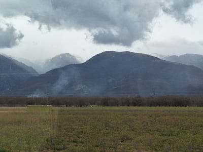 The image depicts an expansive rural landscape in Gabarevo, Bulgaria, under a heavily overcast sky. Dark grey clouds dominate the upper portion of the frame, with lighter grey areas visible between them.
In the foreground, a large field extends across the lower half of the image, covered with low-lying vegetation showing shades of green and brown. A light-colored, rectangular structure, possibly a small building or container, is partially visible within this field, positioned slightly to the left of the central axis, behind a slight rise in the terrain.
The midground features a dense, horizontal line of bare deciduous trees, likely an orchard, stretching across the scene. These trees exhibit visible branches without foliage. Above this tree line, a dark utility pole with visible crossbars and power lines stands slightly left of center. Two distinct plumes of light grey smoke or mist are observed rising from the base of the nearest mountain range, behind the tree line, with the plume on the left being more substantial.
The background consists of multiple layers of mountains. The closest range is dark and appears densely vegetated. Further in the distance, successive mountain ranges are progressively lighter in hue due to atmospheric perspective, with several distant peaks displaying distinct patches of white, indicating snow cover. No individuals or animals are present in the scene.