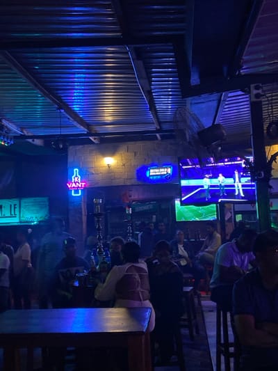 The image depicts the interior of a dimly lit bar or nightclub, located at the coordinates -6.157 latitude, 35.724 longitude, in Dodoma, Tanzania. The space is adorned with neon signs, including "IK VANT" and "EXTRA COLD SINGLE ROOM", both illuminated in a bright blue color. A large television screen displays a sports event, featuring three individuals on the screen, presumably playing a sport. The bar is sparsely populated, with a mix of male and female patrons, some seated at tables while others stand around. Several patrons are engaged in conversations, while others are occupied by various activities like drinking, watching the television, and smoking hookahs. The scene is set in a bar setting with a concrete wall structure, an overhead metal roof, and multiple wooden tables and chairs. The lighting is dominated by an array of bright blue neon lights, casting a vibrant blue hue over the entire space. The overall atmosphere of the scene is relaxed and social, with patrons enjoying a night out.