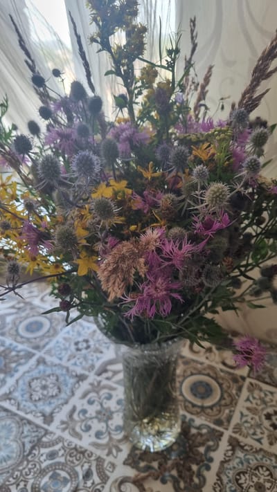 A close-up shot features a bouquet of wildflowers in a glass vase, placed on a table with a tiled pattern. The bouquet is a vibrant mix of purple thistles, yellow wildflowers, and dried grasses. The background is blurred, showing a light-colored curtain and hints of natural light. The overall scene evokes a sense of rustic charm. The image does not provide any specific clues about its location in the city of Aleksin, Russia.