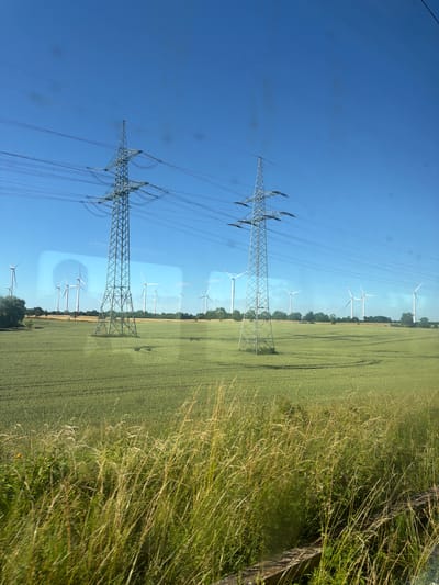 The image depicts a landscape in Angermünde, Germany, characterized by a clear blue sky and a vast green field, likely a crop field, stretching into the distance. The scene is dominated by two large electricity pylons with multiple power lines crisscrossing between them. In the background, several wind turbines are visible, indicating a focus on renewable energy in the area. The field appears to be in daylight, suggesting it is daytime, and the clear sky implies fair weather. The overall impression is one of rural industrialization, where agricultural land coexists with energy infrastructure. There is no visible text in the image.