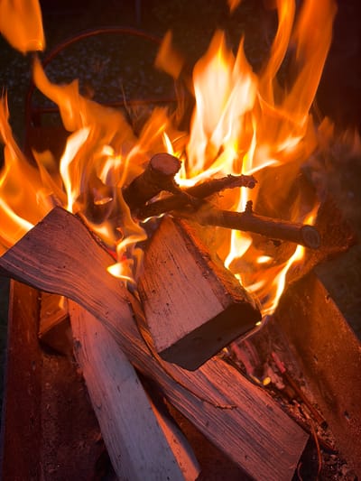 In the city of Манихино Снт, Russia, an outdoor fire pit is ablaze with large, vibrant orange flames. Several pieces of split wood are arranged within the pit, with smaller branches resting atop the larger logs. The wood is actively burning, with some pieces showing glowing embers. The fire pit appears to be made of dark metal. The surrounding environment is dark, suggesting it is nighttime or evening. No people are visible in the image.