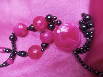 A segment of beaded jewelry is depicted lying on a creased, fuchsia-pink textile surface. The jewelry consists of several large, highly reflective, spherical and oblate beads, approximately 1.5 to 3 centimeters in diameter, exhibiting a marbled pattern of fuchsia and white. These larger beads are interconnected and strung with numerous smaller, spherical beads, approximately 0.5 centimeters in diameter, which appear dark gray with a metallic luster. A visible strand of primarily dark gray beads, interspersed with a few minute translucent beads, extends towards the upper right of the frame. Reflections from an unseen light source are visible on the smooth surfaces of the larger pink beads. No individuals or actions are present in the scene. The location is Qarabucaq qəsəbə, Azerbaijan.