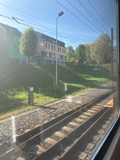 A bright, sunny view from a train window captures the railway line and adjacent landscape in Ettelbruck, Luxembourg. The image, taken at what appears to be morning or late afternoon given the long shadows, shows the train tracks with gravel ballast and concrete sleepers running diagonally across the lower right, receding into the distance.
To the left of the tracks is a lush green grassy verge, bordered by various trees with full foliage, suggesting spring or summer. In the mid-ground on the left, several buildings are visible. One prominent structure is a multi-story building with a light brown facade and numerous windows and balconies, possibly residential or commercial. Below it, a smaller building with a distinctive green facade and a dark roof is partially visible.
Overhead, a clear blue sky spans the upper portion of the frame, crisscrossed by numerous power lines that appear to be part of the railway's electrification system. A tall street lamp post stands prominently in the mid-left. Near the tracks, amidst the grass, a small white marker or signpost is visible with black text. The visible text on this marker reads: "46 A", "95", and "6331". The overall scene depicts a typical European suburban railway environment, bright and clear.