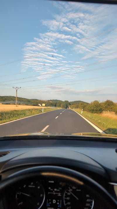 The image depicts a view from the driver's seat of a vehicle, looking out through the windshield at a rural road. The road curves to the right and stretches into the distance. On either side of the road are fields of dry grass and some vegetation, with rolling hills in the background. Power lines are visible stretching across the sky, supported by utility poles. The sky is blue with scattered, wispy clouds. In the foreground, the dashboard and steering wheel of the car are visible, including part of the instrument cluster which shows a gear selection of "D" (Drive). The time of day appears to be late afternoon or early evening, given the lighting. The scene is consistent with a rural landscape in the Czech Republic, potentially near Skršín, given the presence of agricultural fields and rolling hills.