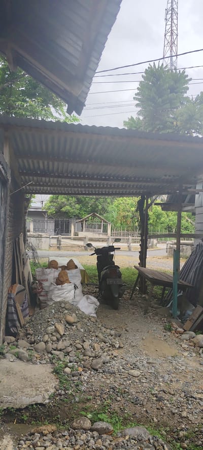 This photograph depicts a utilitarian outdoor area, likely a carport or storage space, in Paloh Teungoh, Indonesia, on an overcast day. A black scooter is parked beneath a corrugated metal roof, which is supported by wooden beams. To the left, several white sacks rest on a pile of rocks and gravel. Some sacks contain what appear to be brown, dried coconut shells or similar agricultural items, and the word "FINASI" is partially visible on one sack. Adjacent to these, various construction materials, including planks, are stacked against a brick wall. On the right, a wooden table is stored against a support post. The ground is uneven, covered with a mixture of dirt, gravel, and rocks, with some small patches of green vegetation.
Beyond the immediate covered area, a glimpse of a residential street is visible, featuring a grey house with a black metal gate and fence. Further back, a small, open-sided wooden structure suggests another storage or market stall. The sky is uniformly grey and cloudy. Overhead, multiple power lines stretch across the frame, and a prominent metal communication tower stands tall against the horizon in the upper right. The scene is devoid of people or active events, conveying a sense of stillness in a working, semi-rural environment.
